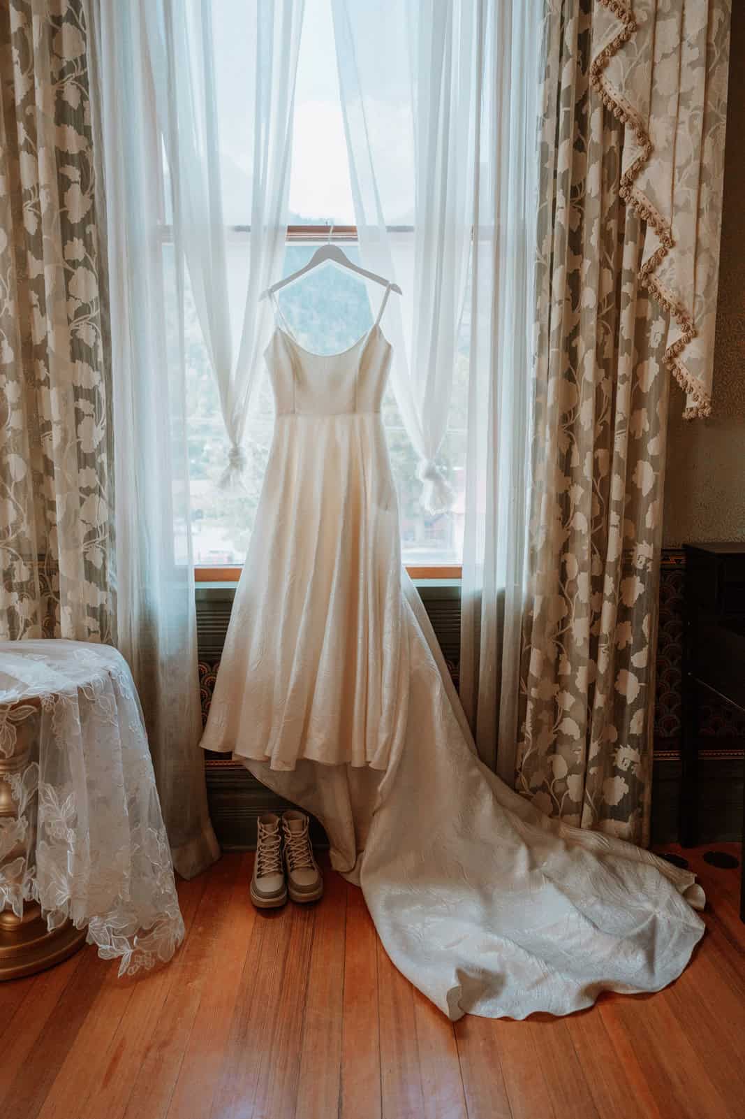 The wedding dress hangs in the window of a vintage Beaumont Hotel room in Ouray, paired with hiking boots below, captured during getting ready photos.