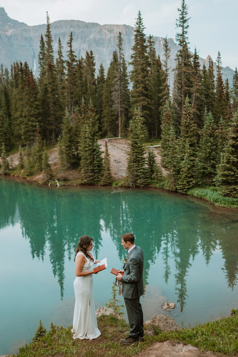 A wide shot of a couple reading their vows from leather books while standing on the grassy edge of a crystal clear turquoise lake reflecting the pine trees during their blue alpine lake vow renewal.