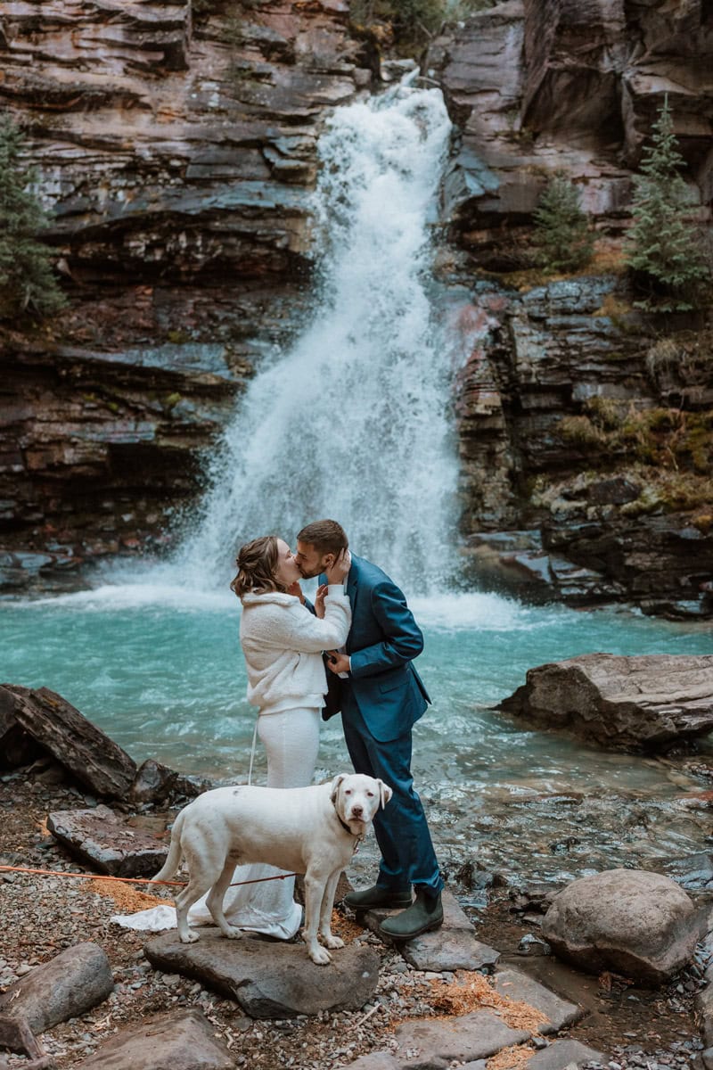 A bride wearing a cozy white jacket and a groom in a blue suit kissing next to a bright blue waterfall pool with their white dog during their Silverton elopement in the San Juan Mountains.