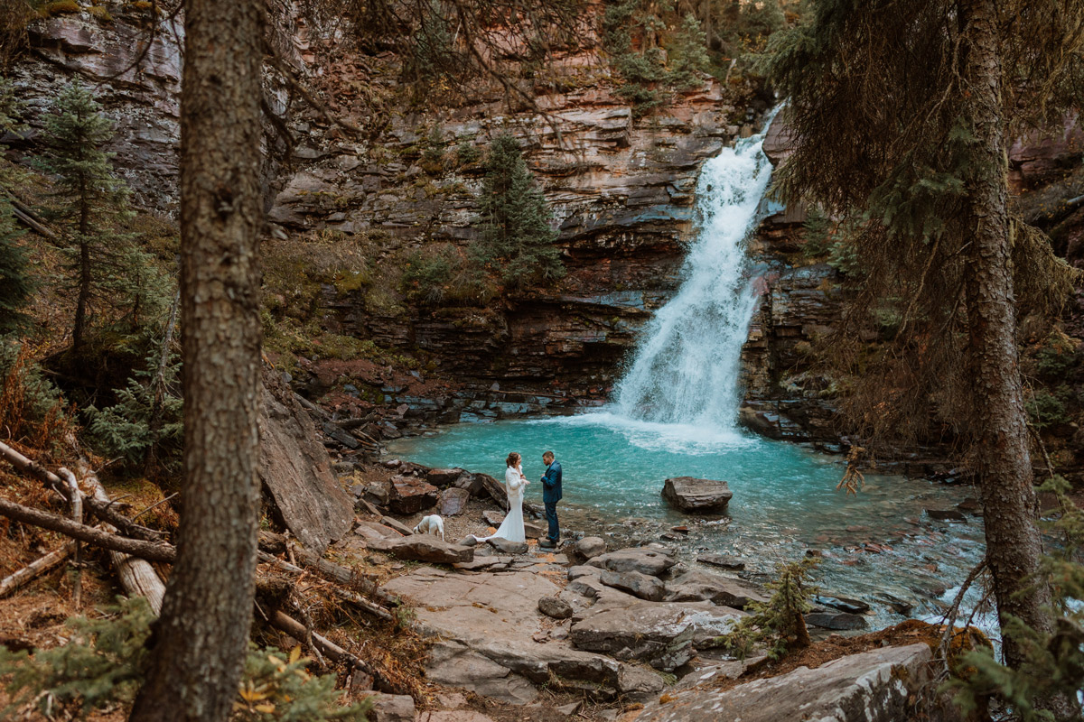 A couple and their dog standing at the base of a rushing turquoise waterfall surrounded by red rocks and pine trees during their intimate blue waterfall Silverton vow renewal ceremony.