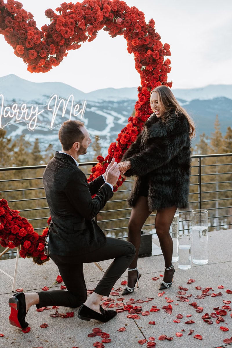 A luxury proposal in Breckenridge, Colorado, featuring a man on one knee in front of a giant heart-shaped red rose installation and a neon "Marry Me!" sign.