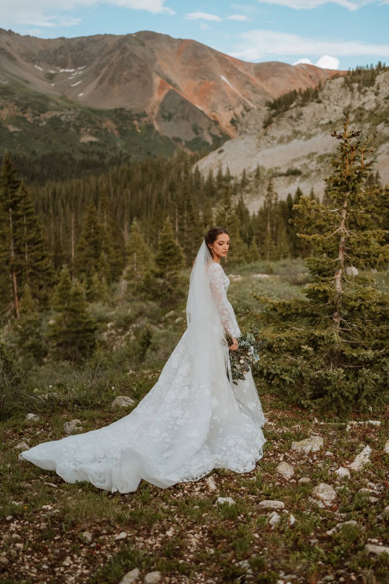A bride poses for a bridal portrait in the mountains for her Aspen elopement in Colorado. The vast mountains rise behind her and her trail from her wedding dress is spread out.
