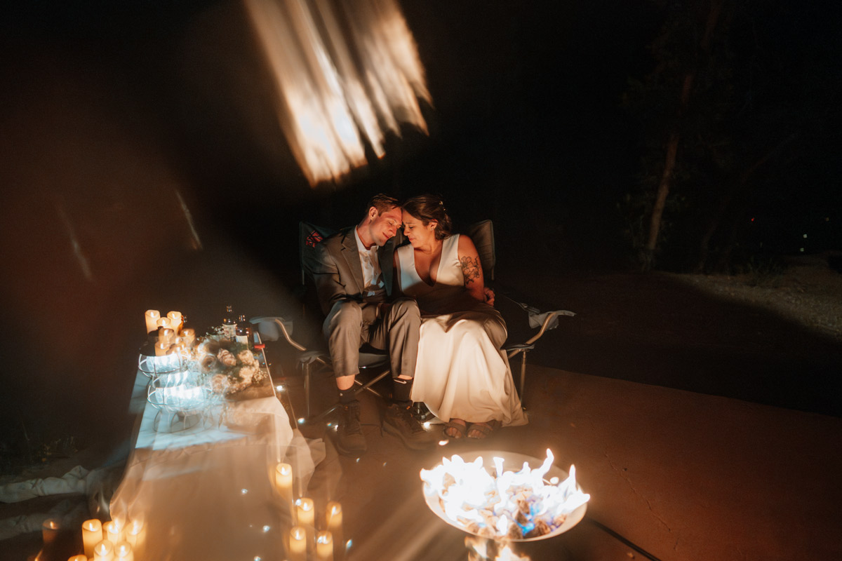A couple cuddling in camping chairs next to a glowing fire pit and a candlelit table with cake and drinks during their romantic night time campfire picnic wedding vow renewal after-party.