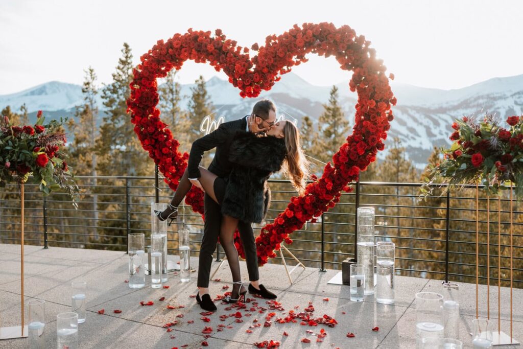 A couple celebrates with a dip kiss in front of an epic red rose heart arch during a Colorado luxury proposal in Breckenridge.