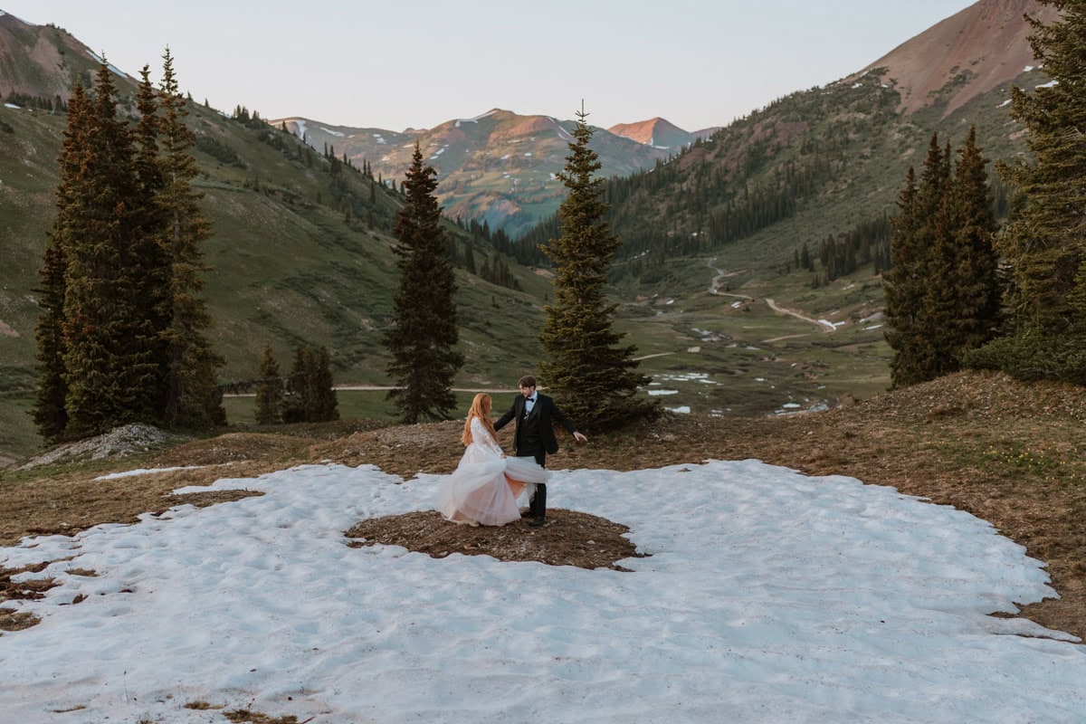 A bride and groom dancing and twirling on a lingering patch of snow high in the mountains surrounded by green pines during their adventurous Colorado wedding vow renewal.