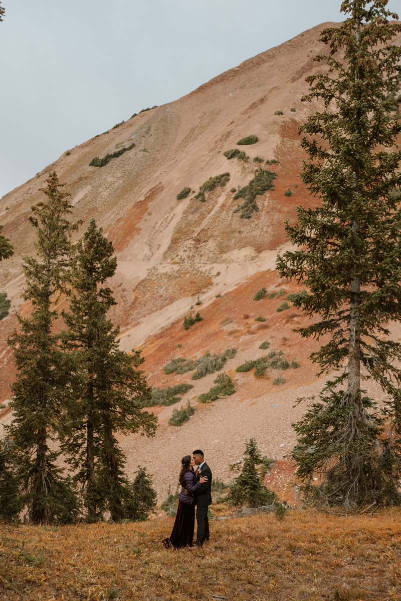 A couple sharing a romantic kiss in an autumn meadow with the bride wearing a purple puffy jacket in front of a massive red mountain slope during their Crested Butte wedding vow renewal.