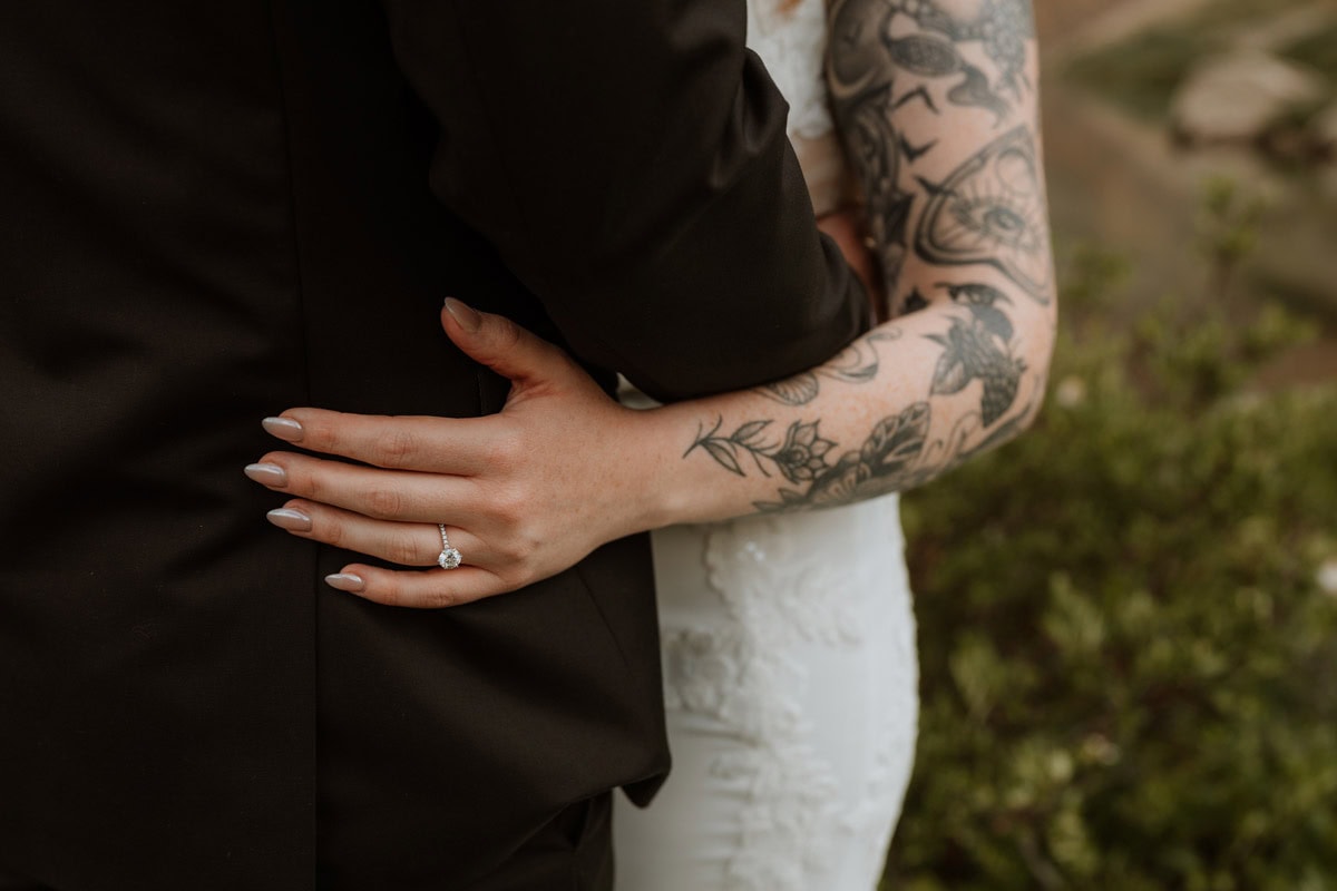 Close up of a bride's hand with a diamond wedding ring resting on the groom's back, showcasing her detailed arm tattoos.