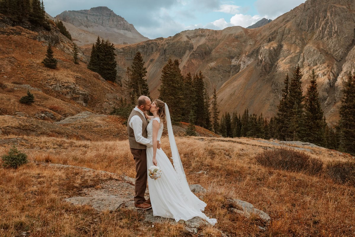 A bride and groom sharing a kiss while standing on a rock among golden autumn grass and pine trees during their fall elopement photos near Ouray in the San Juan Mountains.