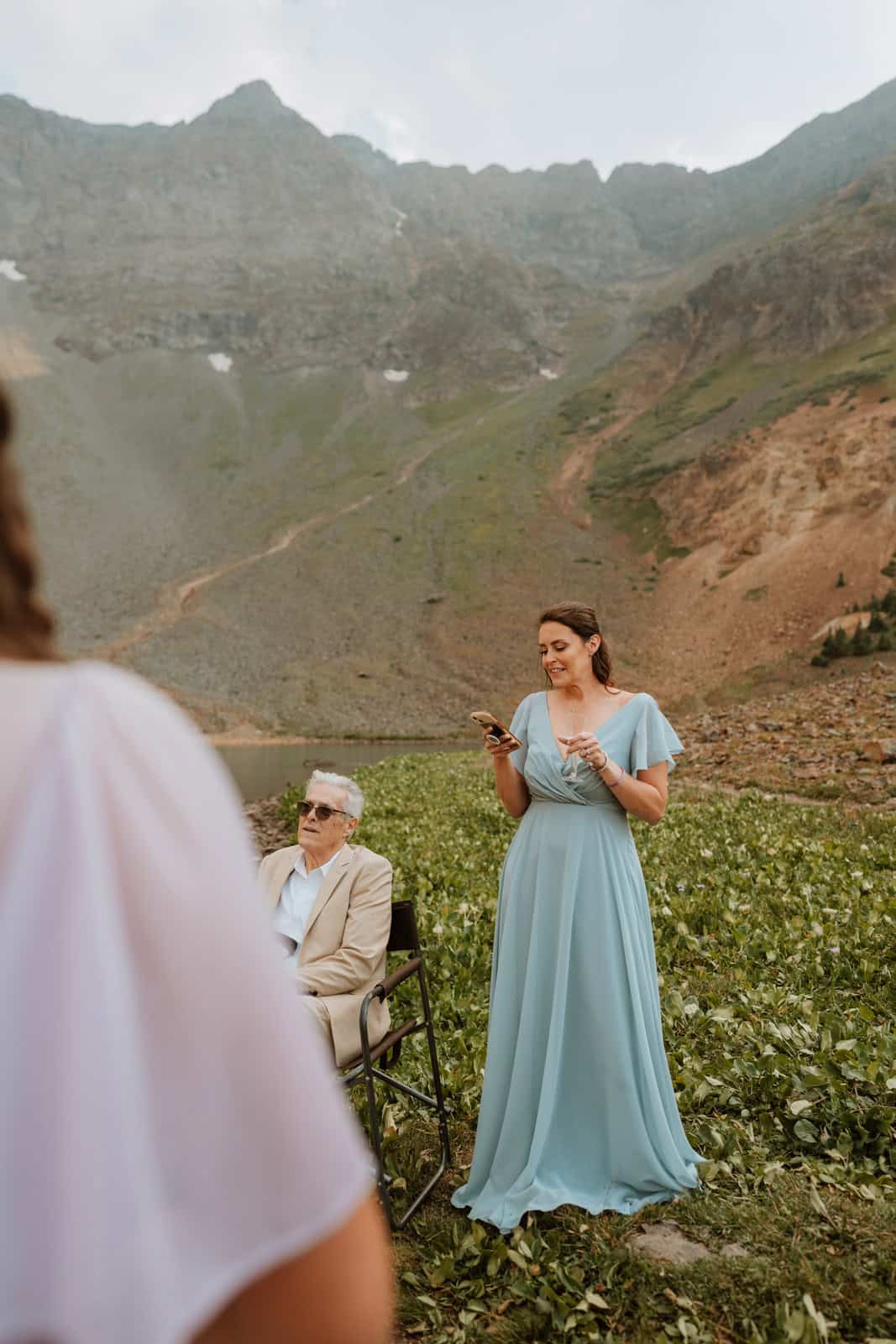 A bridesmaid in a soft blue dress delivers emotional wedding speeches during an intimate elopement ceremony in the San Juan Mountains, with a seated guest and dramatic alpine scenery in the background.