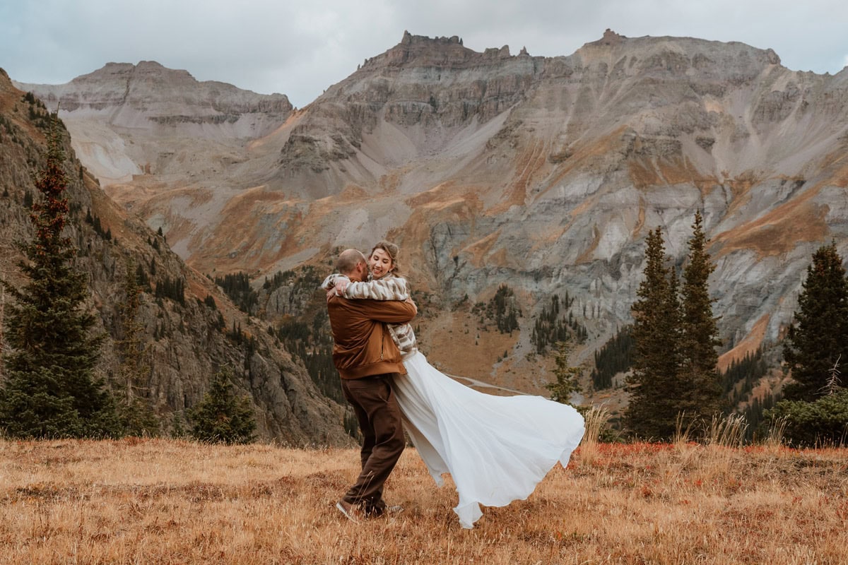 A groom playfully lifting and spinning his bride in a grassy alpine meadow during their fall Ouray elopement, surrounded by the dramatic, rugged peaks of the San Juan Mountains.