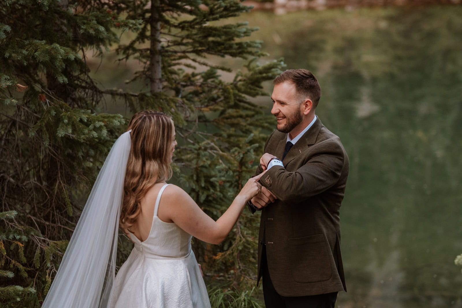 A joyful groom smiles as he looks at his bride for the first time, while she checks out his custom cuff links. The couple stands beneath the trees, sharing a tender and playful moment next to a mountain lake.