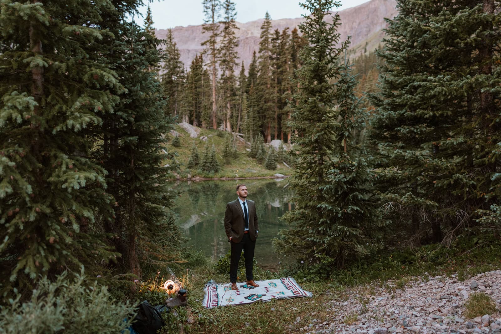The groom stands confidently on a patterned rug laid out in the forest, adjusting his suit and soaking in the scenery before the first look by an alpine lake in the San Juan Mountains.