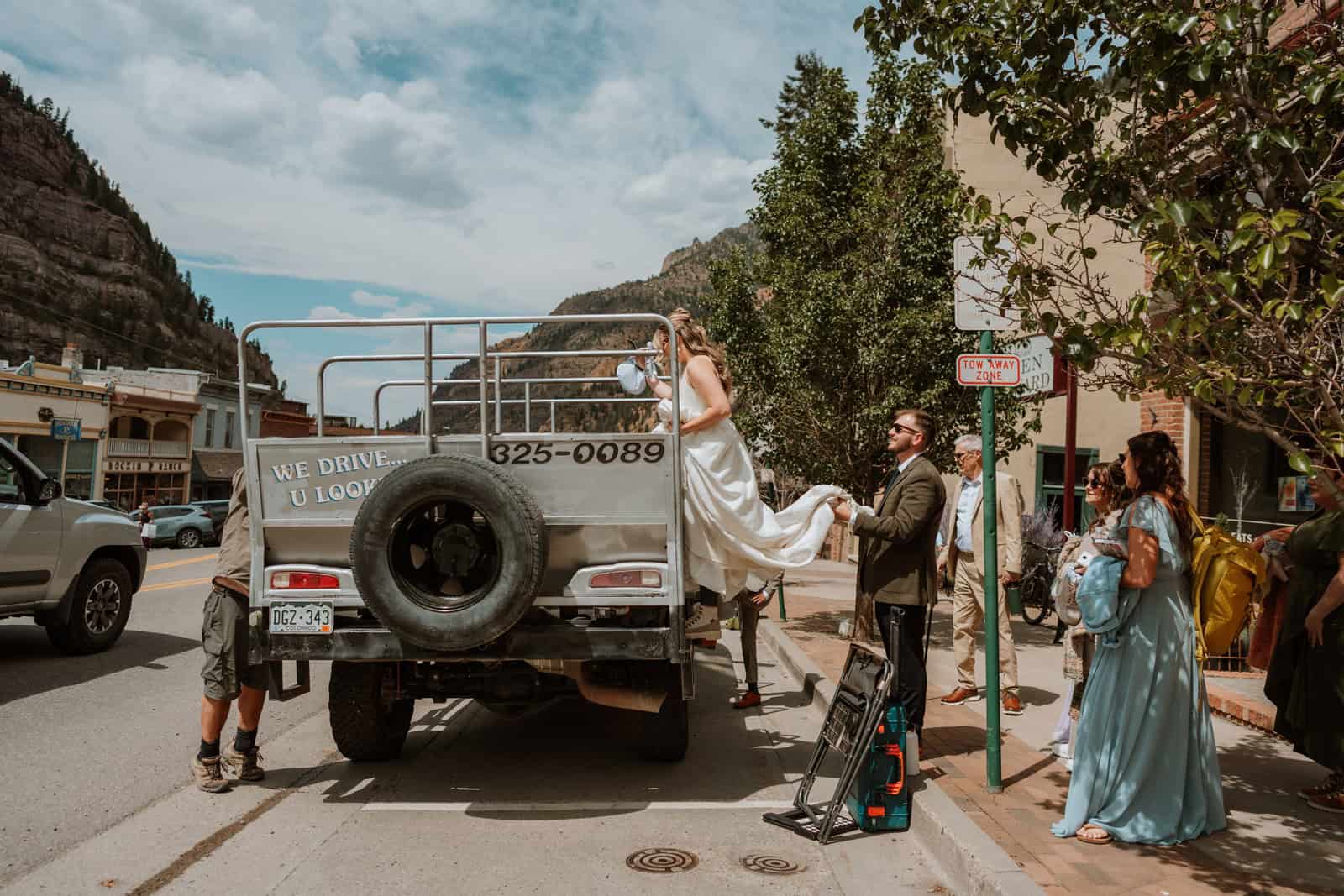 A bride in her wedding dress steps into the back of a rugged tour jeep in downtown Ouray, while the groom and a small group of guests help and watch, ready for their wedding jeep tour adventure.