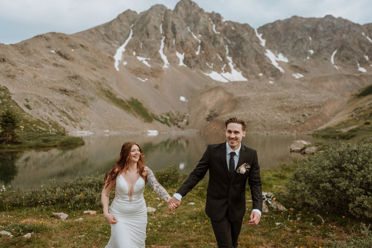 A joyful bride and groom holding hands and walking through a mountain meadow after their Aspen elopement ceremony.