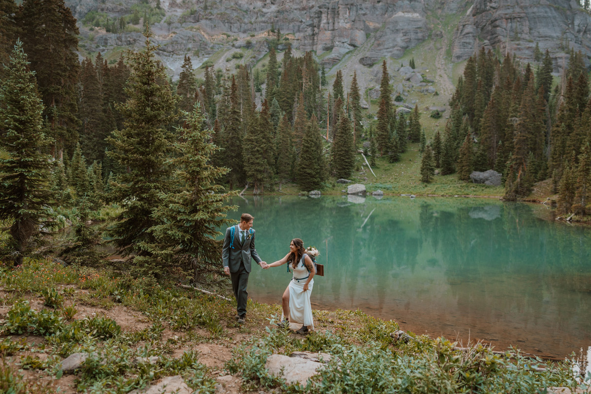 A bride and groom wearing hiking backpacks walking up a trail away from a stunning turquoise alpine lake surrounded by pine trees during their adventurous wedding vow renewal.