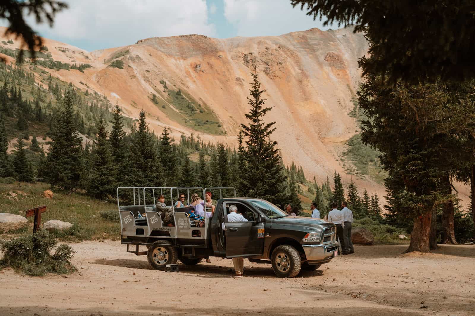 A group jeep tour parked in a scenic forest clearing near Ouray, surrounded by pine trees and bright orange rocky mountains in the distance.