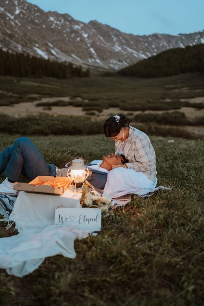 A romantic shot of a groom resting his head in his bride's lap on a mountain meadow blanket at dusk, enjoying a casual pizza picnic vow renewal Colorado style dinner with a glowing lantern and a We Eloped sign.