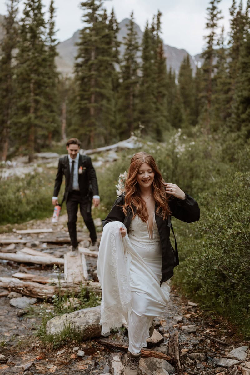 A bride lifting her white wedding dress to navigate a log crossing over a stream during her hike down the mountain.