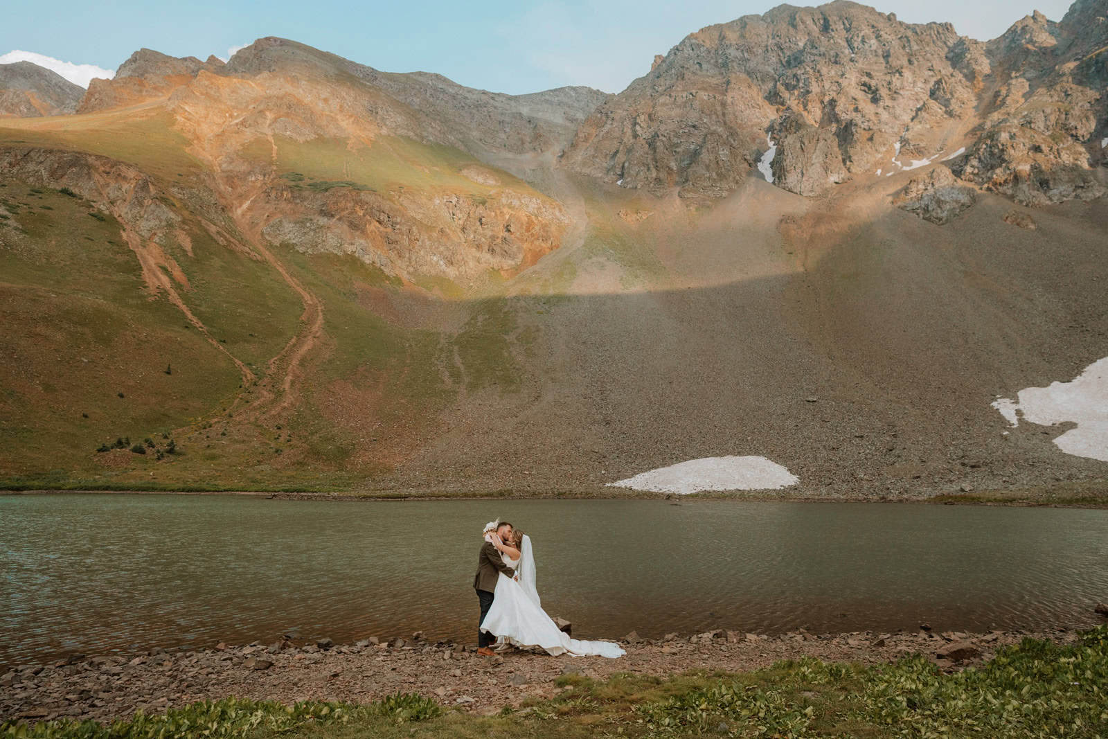 A wedding couple shares a kiss while standing on the rocky shore of a mountain lake. The bride's long white veil flows behind her as she embraces the groom. The large, sun-dappled mountain from the previous images serves as the backdrop.