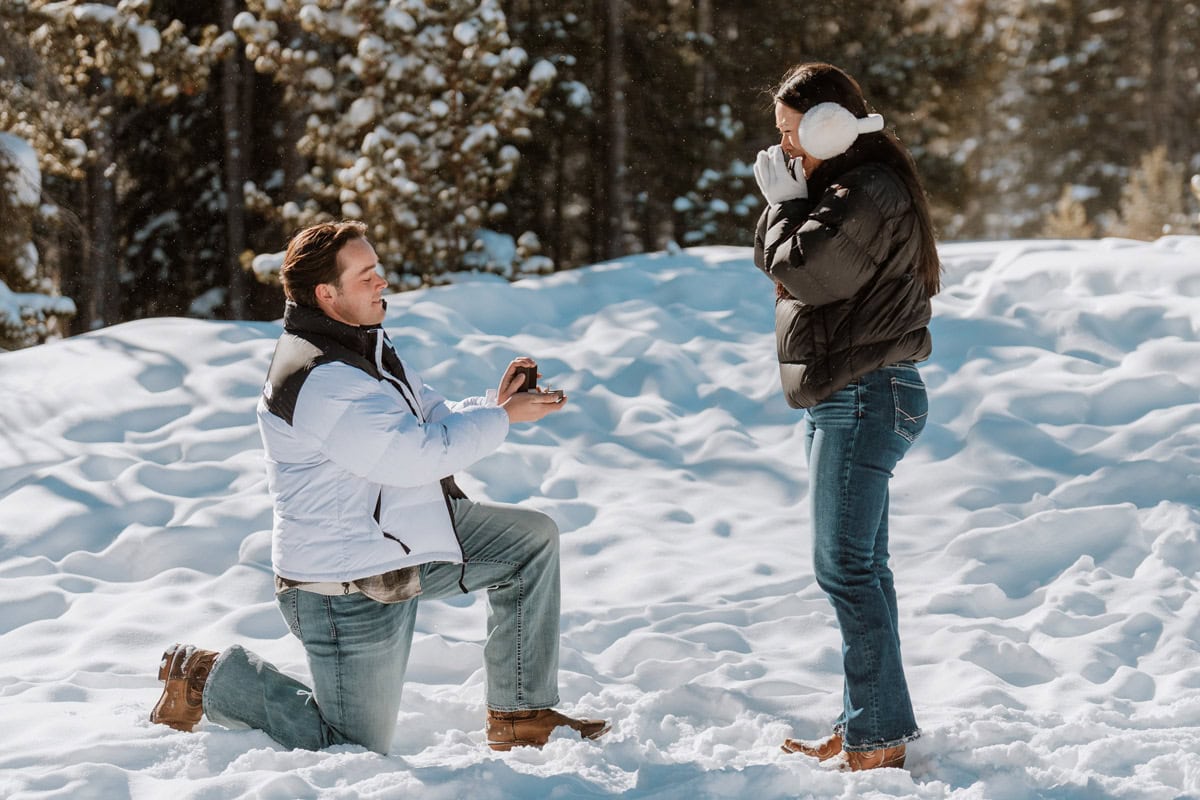 A man in a winter vest proposes on one knee to his partner in a snowy forest during a Colorado luxury proposal.
