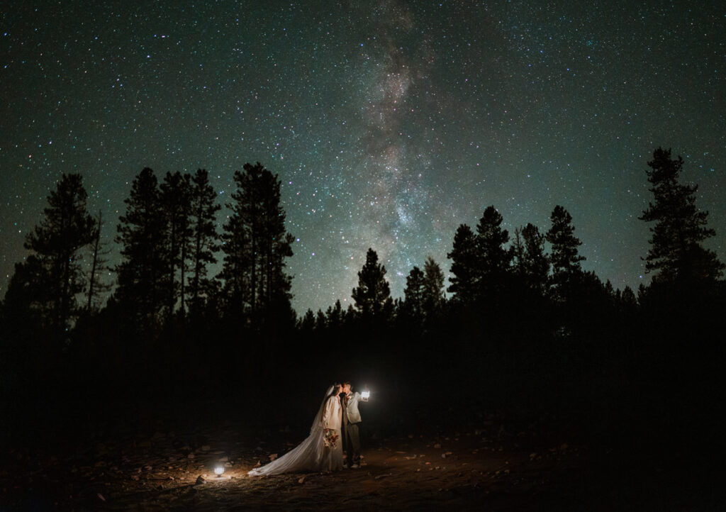 A breathtaking night sky shot showing the Milky Way galaxy stretching over a silhouette of pine trees while a couple kisses holding a glowing lantern during their star photos wedding vow renewal.