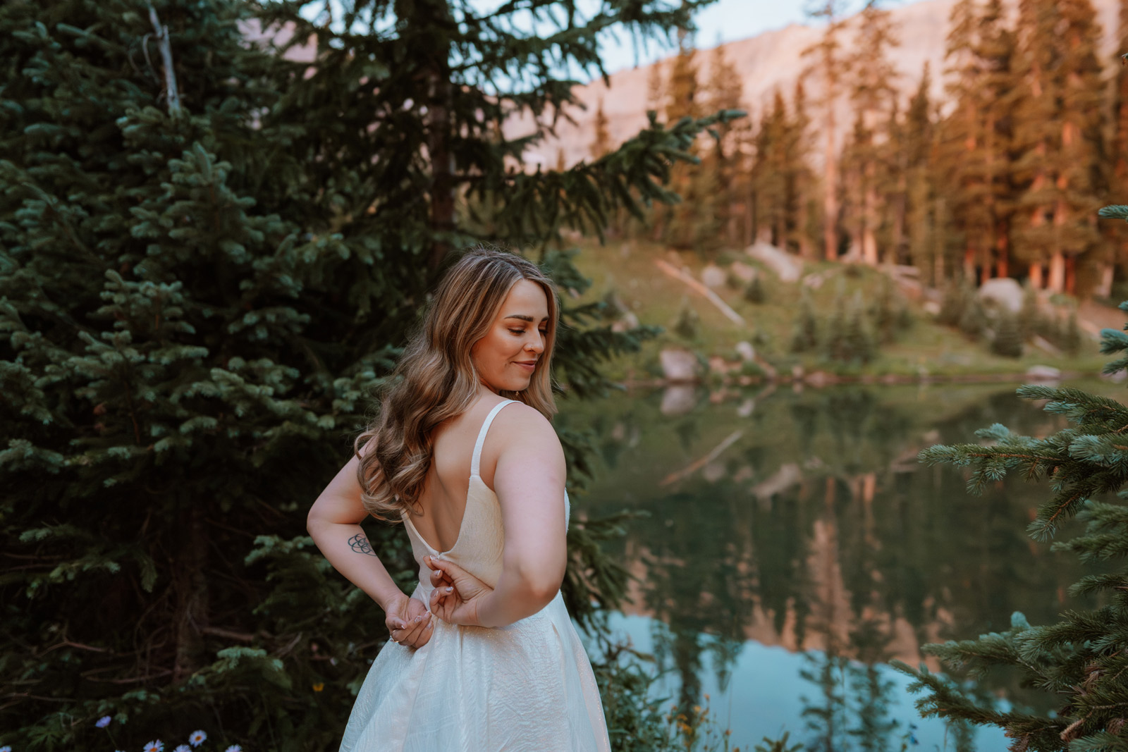 Bride smiles while holding her wedding dress during sunrise getting ready elopement photos in the forest near a mountain lake.