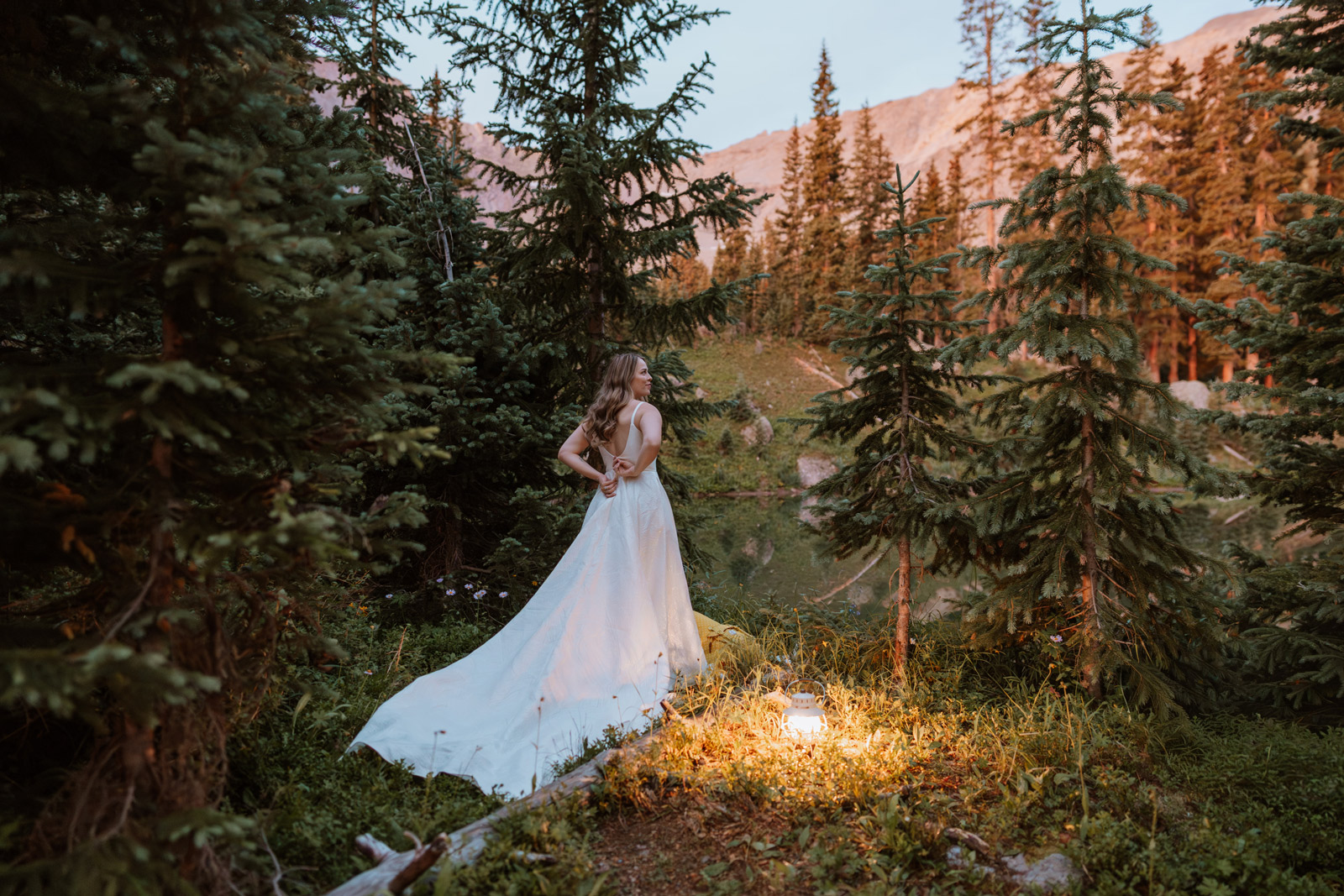 Bride stands in the forest at dawn wearing her wedding dress, backlit by sunrise light during getting ready photos for a San Juan Mountains elopement.