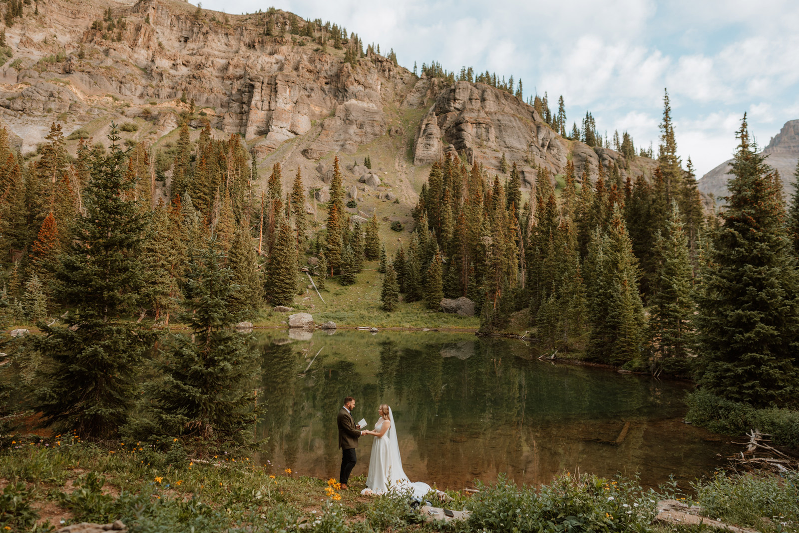 Bride and groom exchange vows at sunrise beside a clear alpine lake surrounded by evergreens in the San Juan Mountains near Ouray during their elopement.