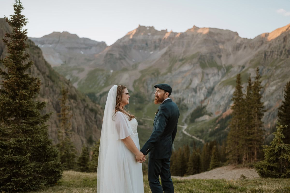A bride and groom holding hands and smiling at each other during their sunset 4x4 elopement near Ouray, Colorado, with towering San Juan Mountain peaks and evergreen trees in the background.