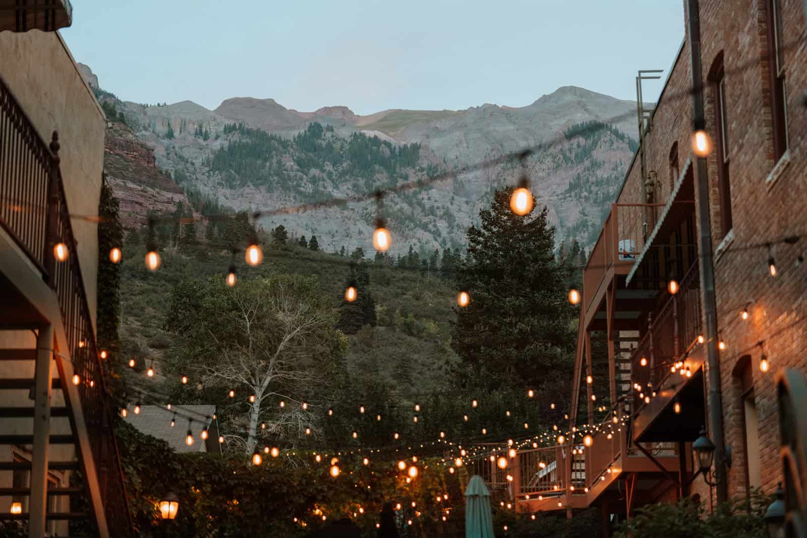 String lights glowing against a dusk sky during a sunset wedding at The Beaumont in Ouray, framed by historic brick buildings and mountain peaks.