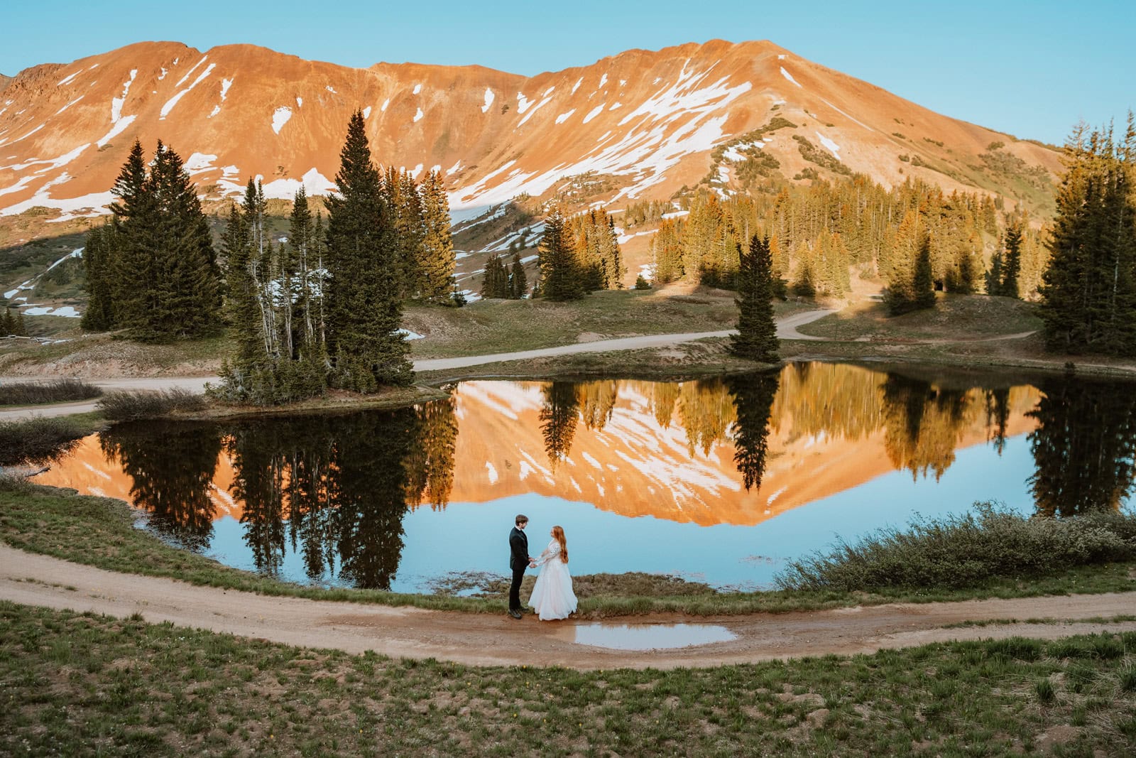 A wide scenic shot of a couple holding hands by a reflective alpine lake with a massive orange mountain peak glowing behind them during their sunset wedding vow renewal ceremony in the Colorado mountains.