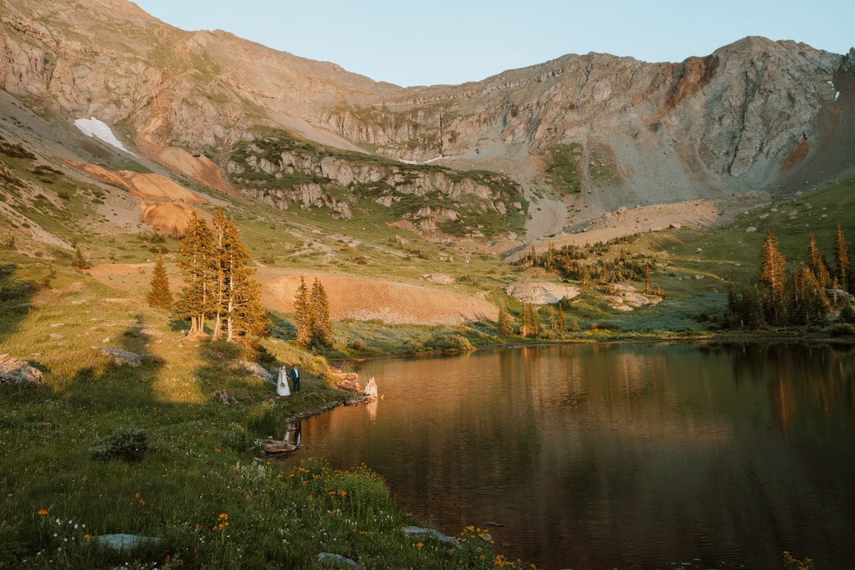 A bride and groom walking along the grassy shoreline of a calm alpine lake at sunset during their Telluride summer elopement.