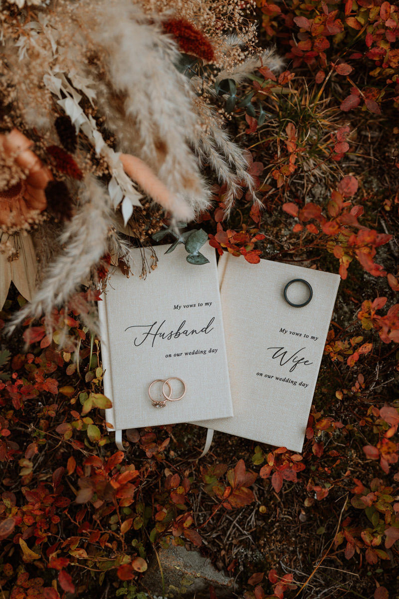 A detail shot of vow books titled "My vows to my Husband" and "My vows to my Wife" resting on autumn leaves next to wedding rings for a wedding vow renewal ceremony.