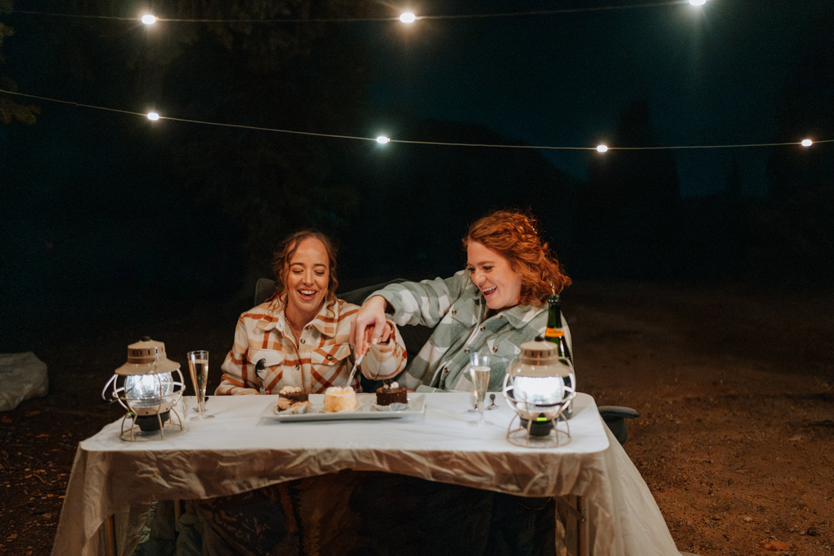 Two brides laughing while cutting a small cake at a private camping table under string lights and lanterns during their cozy outdoor vow renewal celebration in the woods.