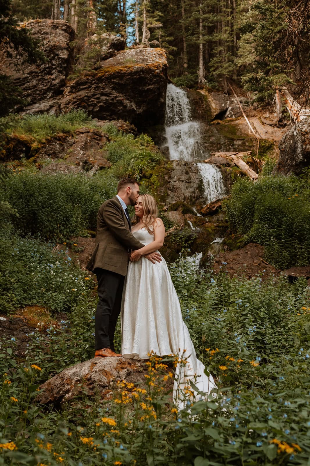 The couple stands on a rock surrounded by wildflowers beneath a cascading waterfall in Ouray, sharing a quiet embrace during their wedding portraits.