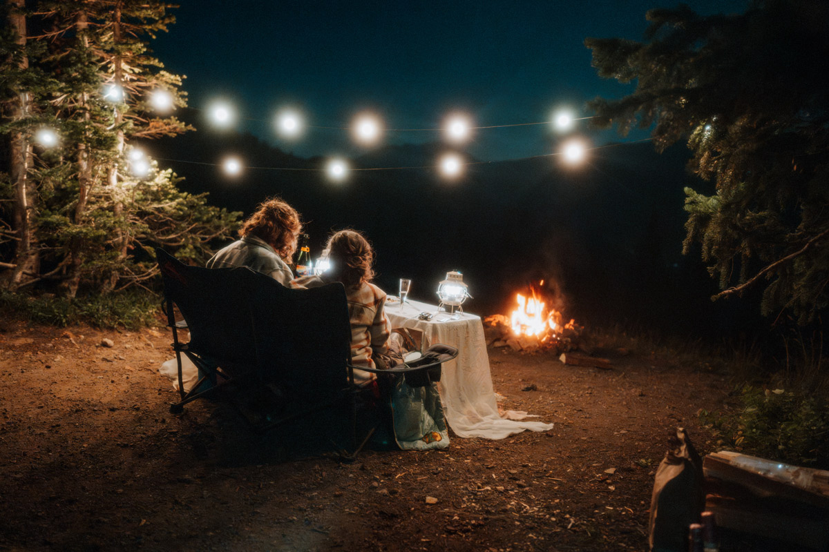 A couple sitting in camping chairs with their backs to the camera, enjoying a cozy fire and string lights at a table set for two during their intimate wedding vow renewal campfire picnic in the woods.