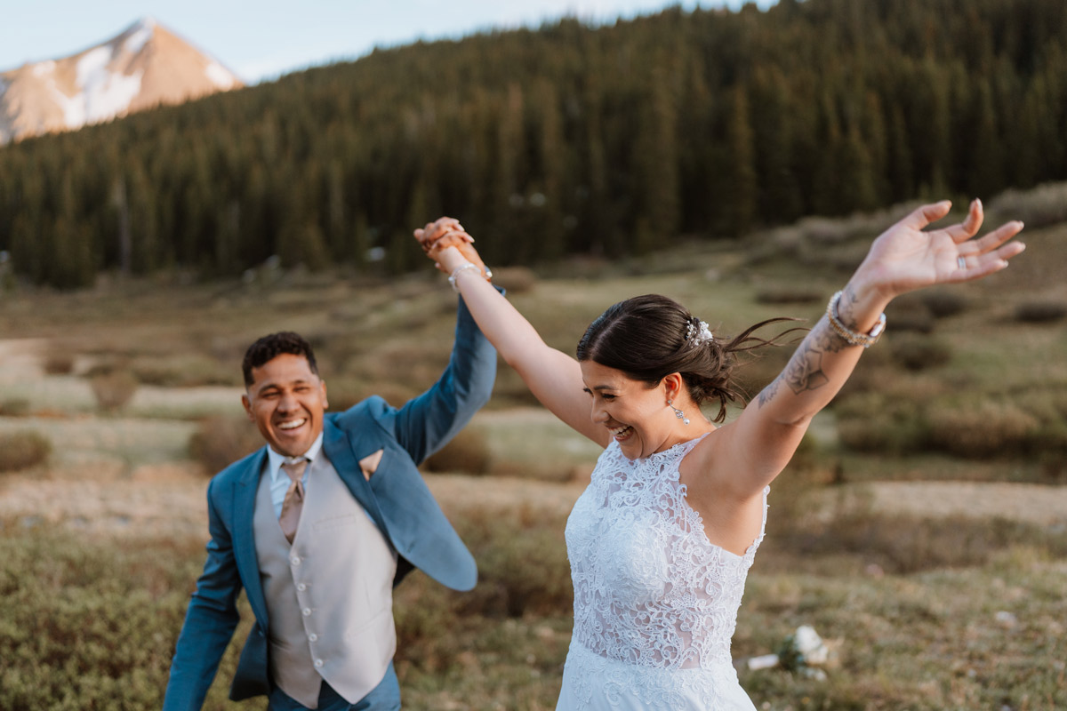 A close-up of a joyous bride and groom throwing their hands in the air and laughing excitedly in a mountain meadow during their wedding vow renewal ceremony celebration.