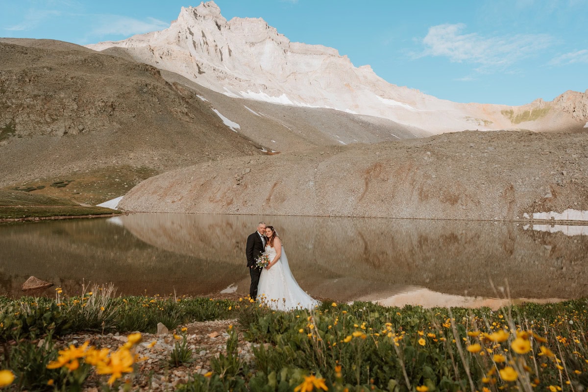 A bride and groom standing by the calm waters of an alpine lake reflecting towering mountain peaks, with bright yellow blooms in the foreground during their wildflower portrait for their Ouray elopement.