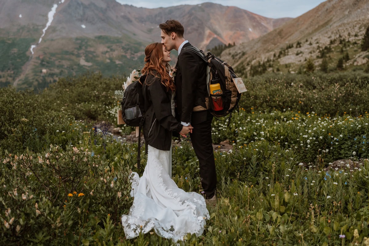 Groom kissing a bride's forehead in a vast mountain meadow filled with white and yellow wildflowers during this Aspen elopement.