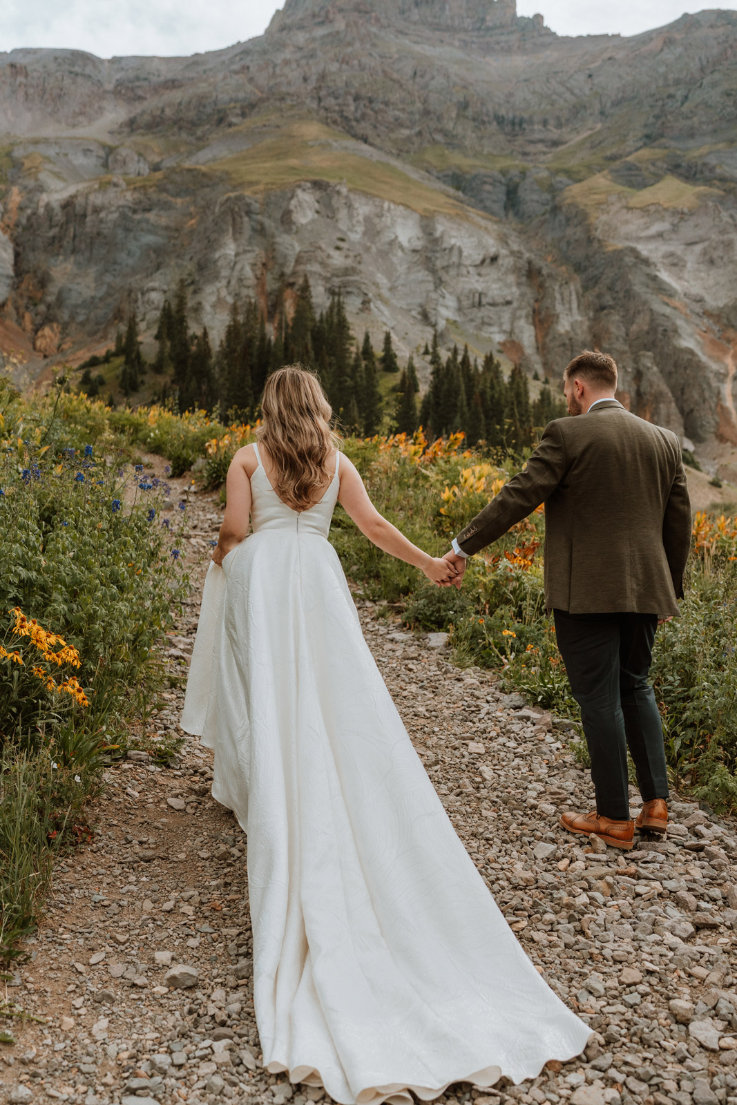 Bride and groom walk hand-in-hand along a wildflower-lined mountain path during their San Juan Mountains elopement.
