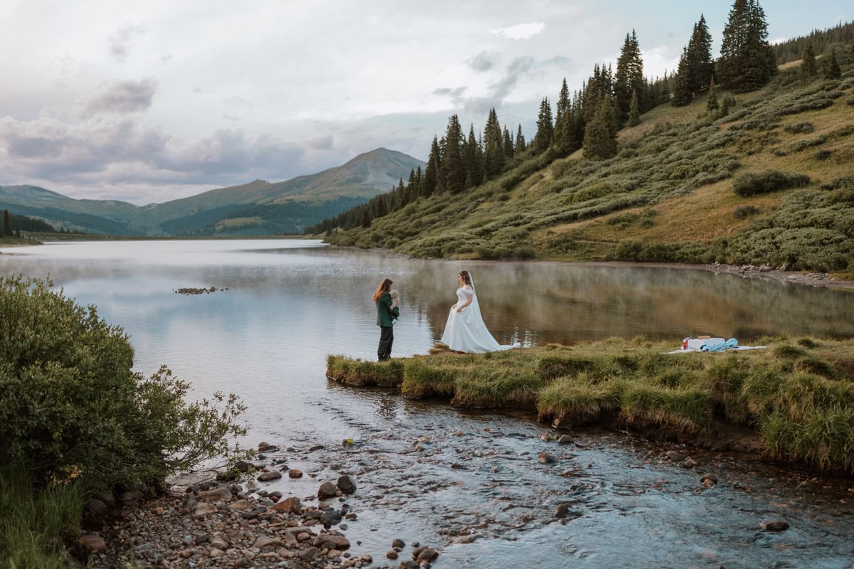 A couple enjoying a private picnic next to an alpine lake after their adventurous mountain destination vow renewal in Colorado. They both walk out to a piece of land extending into the lake with the picnic to the right of the image.