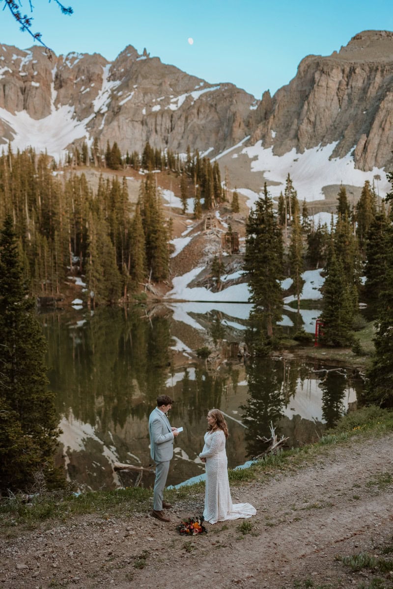 A groom reading his promises from a vow book to his bride standing beside a calm reflective lake with rugged mountain peaks in the background during their Alta Lakes destination vow renewal.