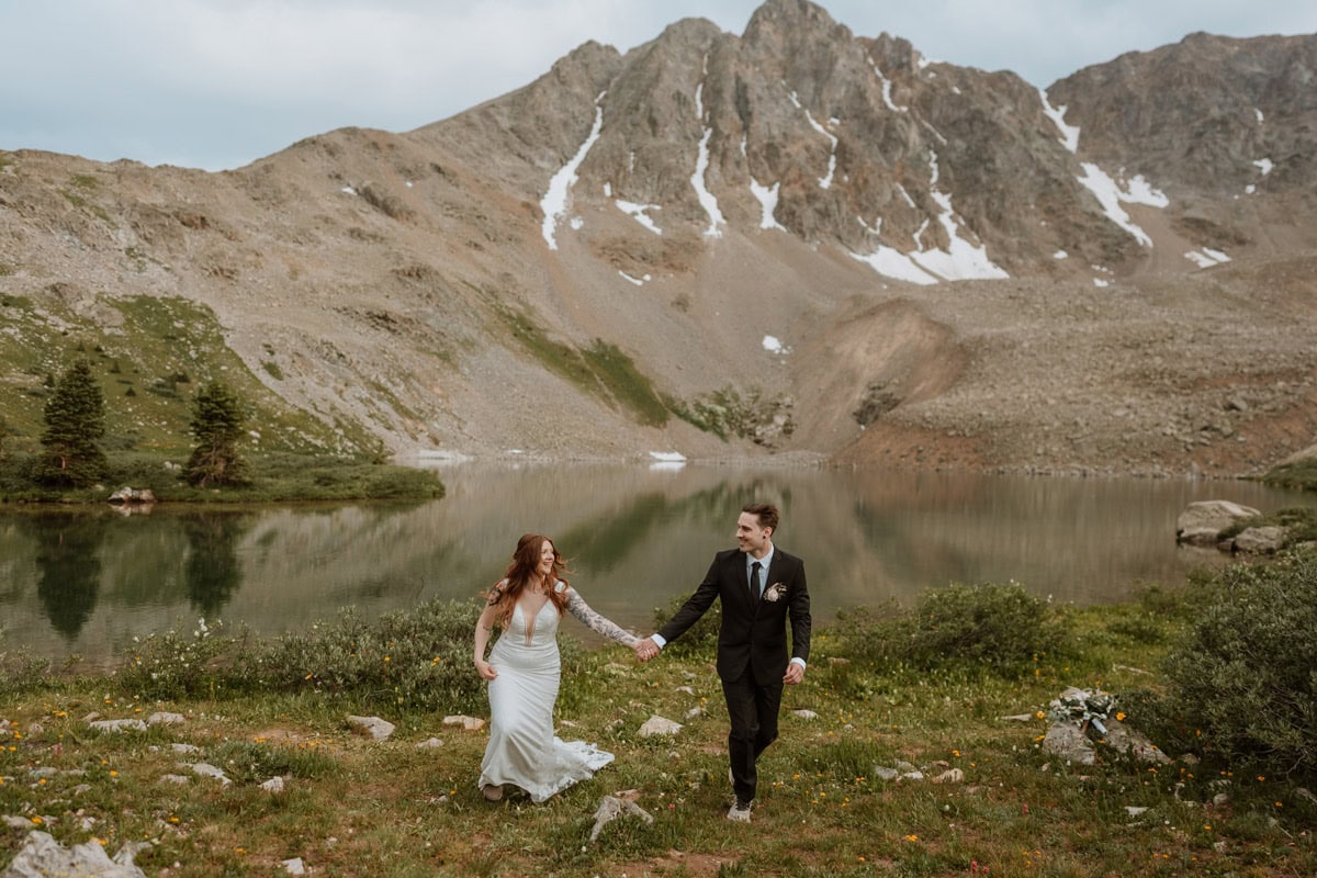 A happy couple walking hand in hand and laughing next to a crystal clear mountain lake during their summer destination vow renewal in Aspen, Colorado.