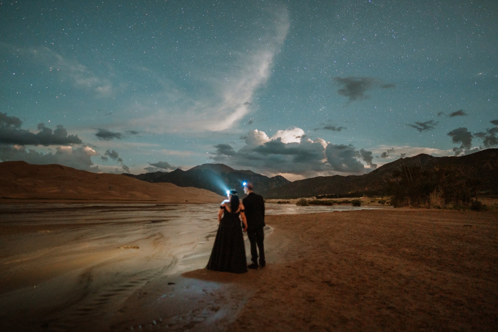 A bride in a black wedding dress and a groom hold hands and look out at the stars during their astrophotography elopement photos at the Great Sand Dunes National Park.