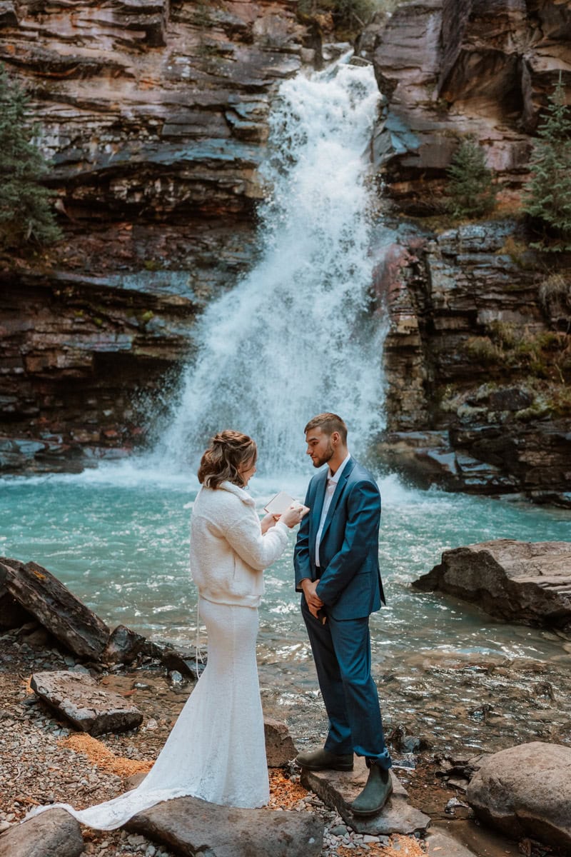 A groom in a blue suit listens warmly as his wife reads her marriage renewal vows by a rushing blue waterfall in a rocky canyon in Colorado.