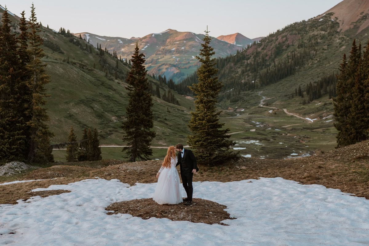 A couple sharing a kiss while standing on a lingering patch of greenery surrounded by white snow in a beautiful green mountain valley during their June destination vow renewal in Crested Butte.