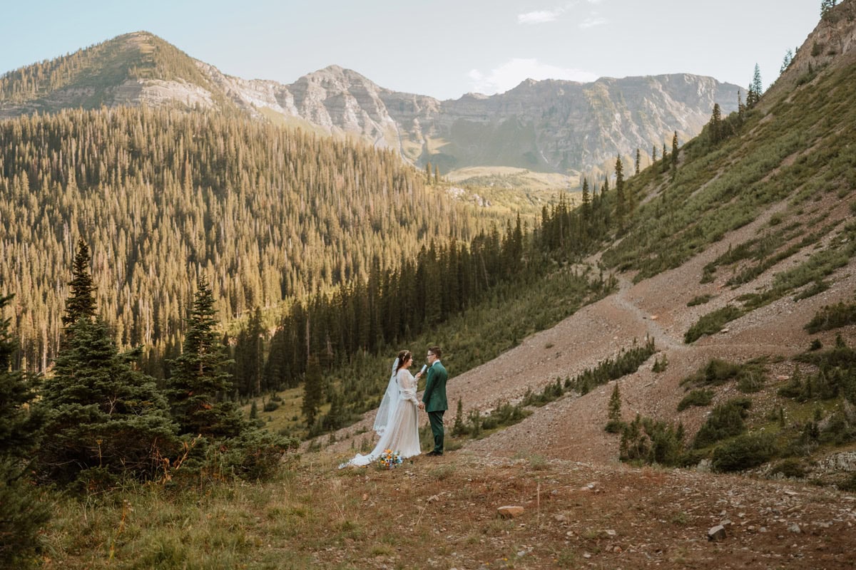 A couple holds hands and reads their vow renewal vows in a sweeping green mountain valley with tall pine trees in Crested Butte.