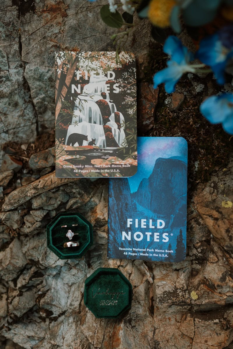 Two national park themed Field Notes custom marriage renewal vow books resting on rocks next to a dark green velvet ring box.
