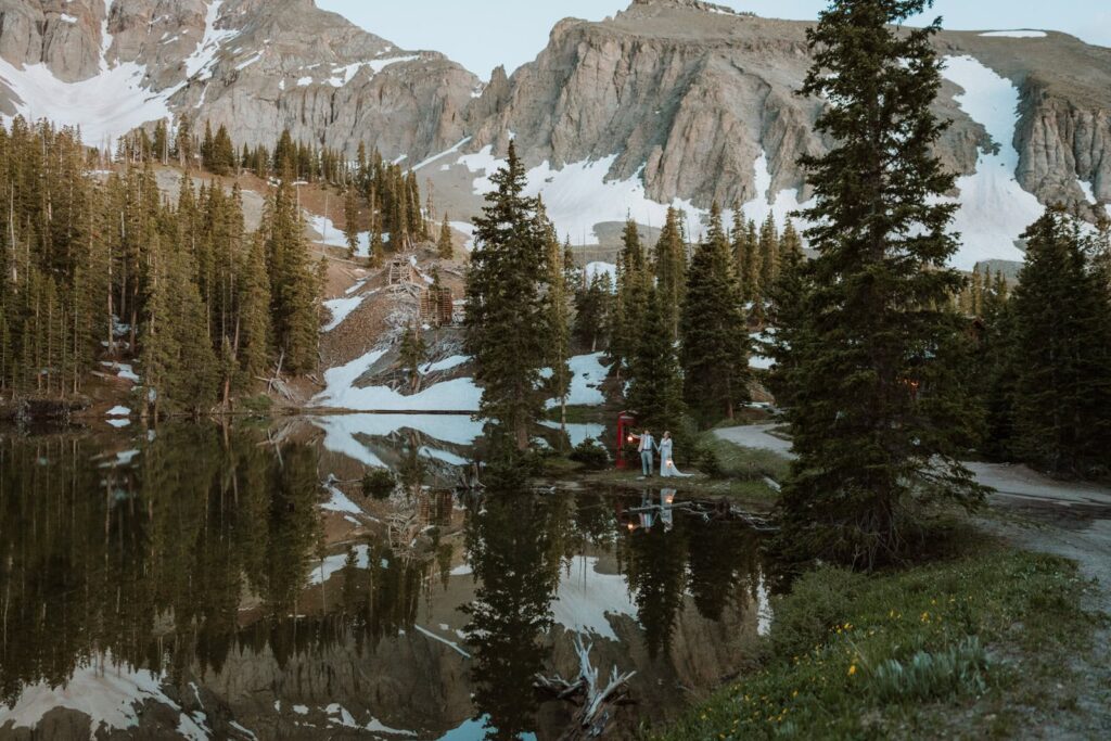 A wide view of a couple standing next to a perfectly reflective alpine lake surrounded by evergreen trees and snow capped mountains during their destination vow renewal at Alta Lakes.