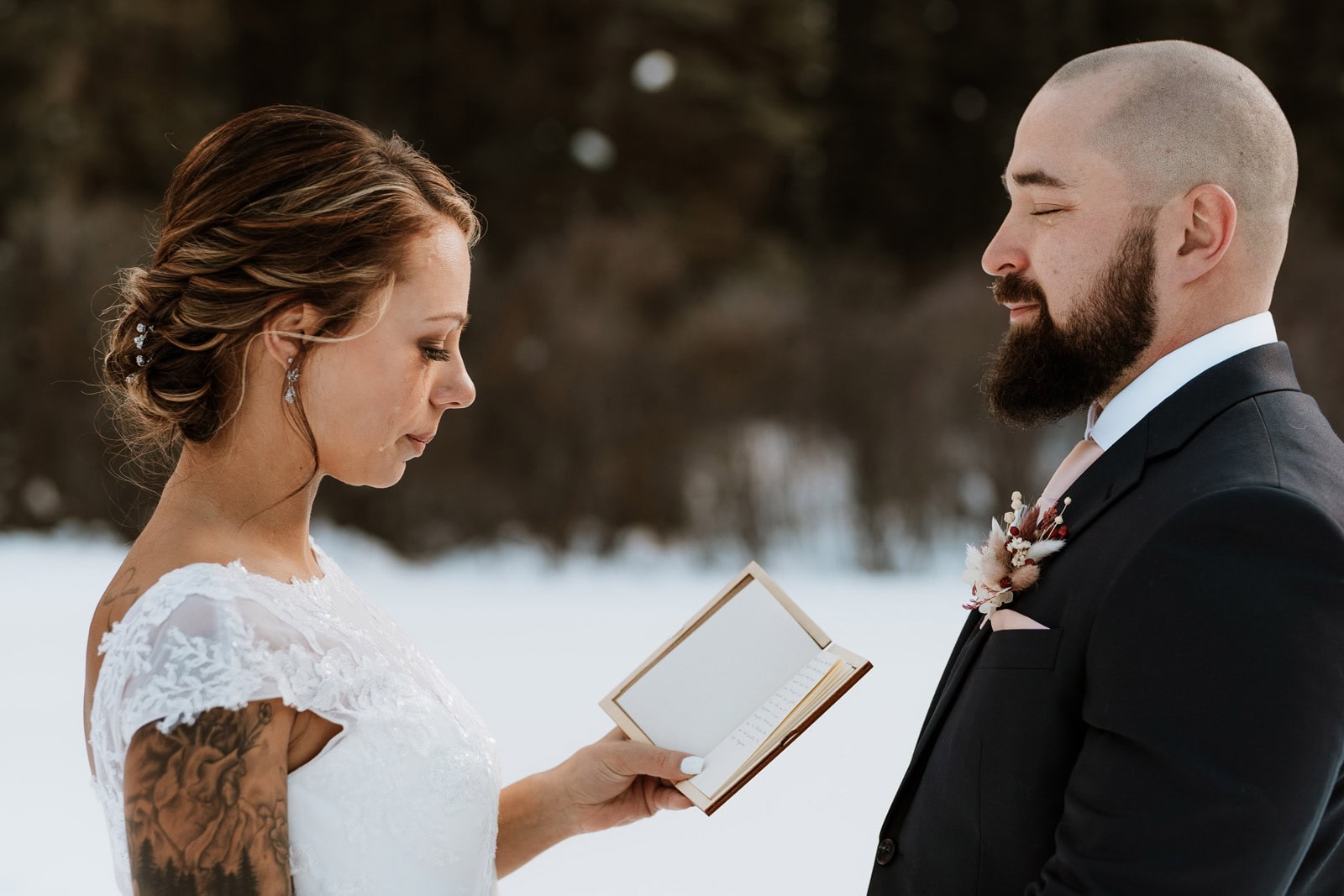 A bride with a sleeve arm tattoo shedding tears while reading emotional vow renewal vows to her husband in a snowy winter landscape.